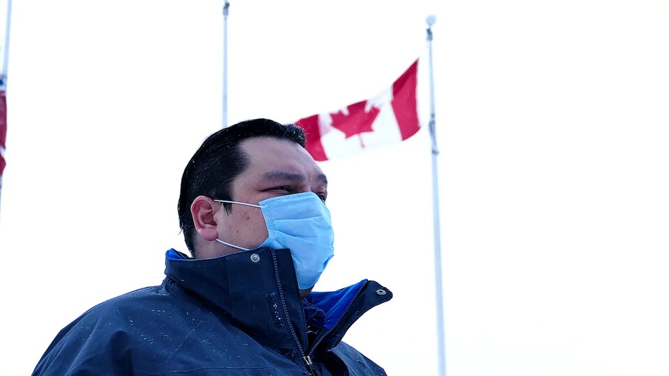 Eric Redhead est dehors devant un drapeau du Canada. Il est vêtu d’un manteau et porte un masque.