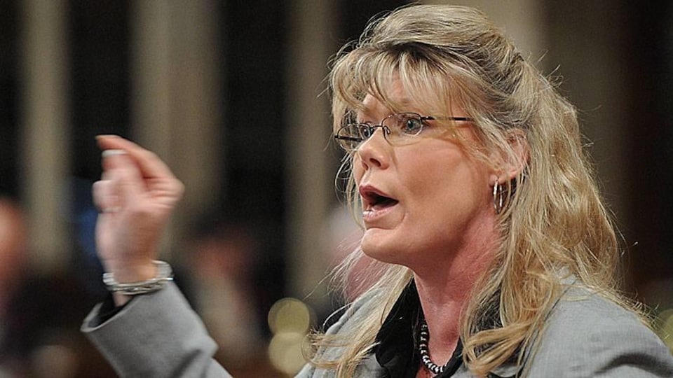 Une femme blonde à lunettes parle en gesticulant.