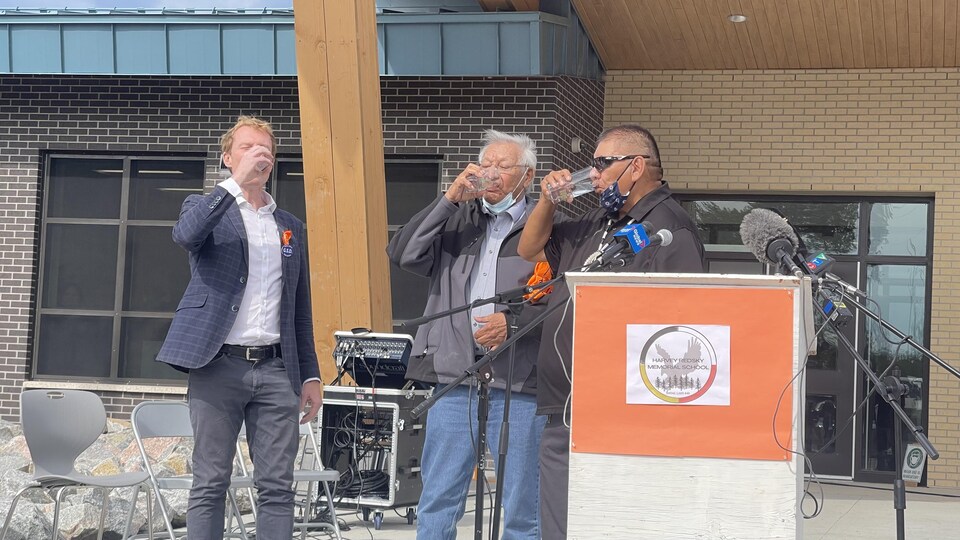 Trois hommes devant un lutrin boivent de l'eau.