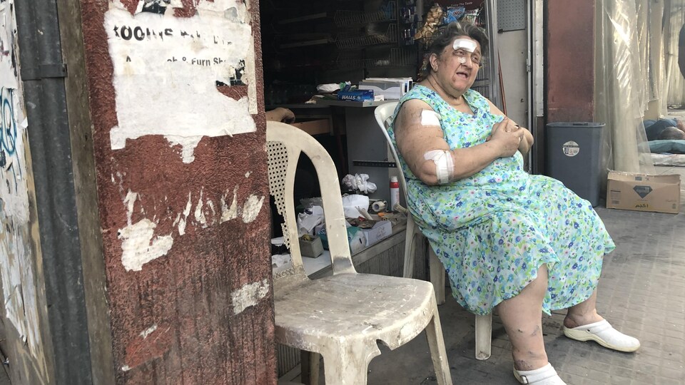 Une femme, couverte de bandages, est assise sur une chaise usée devant un commerce en désordre.