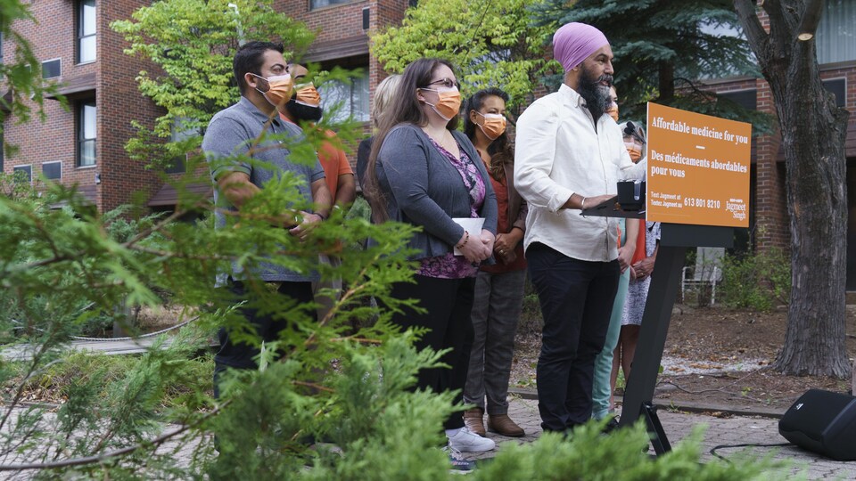 M. Singh est de profil et il parle aux représentants des médias. Devant lui, on peut lire « Des médicaments abordables pour vous ». Autour de lui se trouvent des candidats du Nouveau Parti démocratique. Le point de presse se déroule dans une allée piétonne entre des immeubles résidentiels.