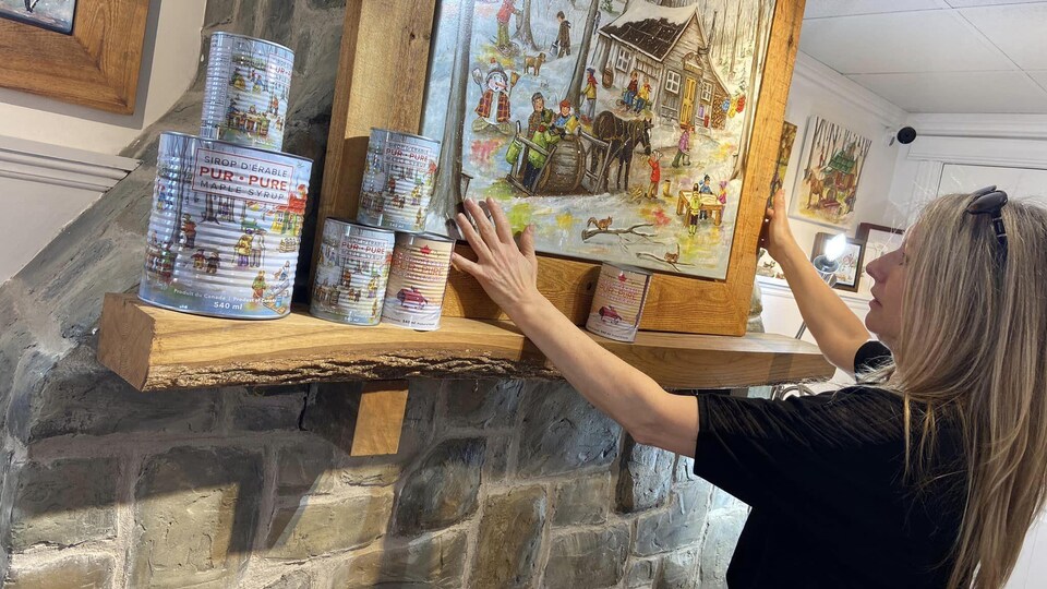 Christine Genest est en train d'ajuster une toile posée au-dessus d'une cheminée. Des boites de conserve de sirop peintes sont également posées sur une planche de bois.