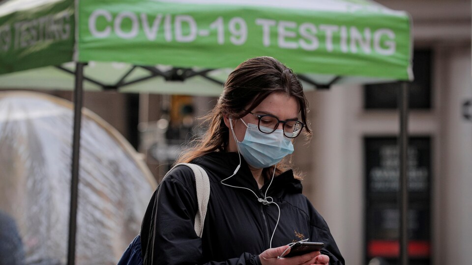 Une femme dans la rue portant un masque et jetant un regard sur son téléphone. Elle passe sous un chapiteau vert sur lequel est écrit « Covid Testing ».