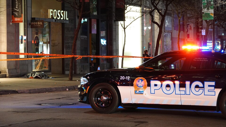 Un véhicule du Service de police de la Ville de Montréal au premier plan et un corps d'un corps recouvert d'une couverture plus loin sur le trottoir.