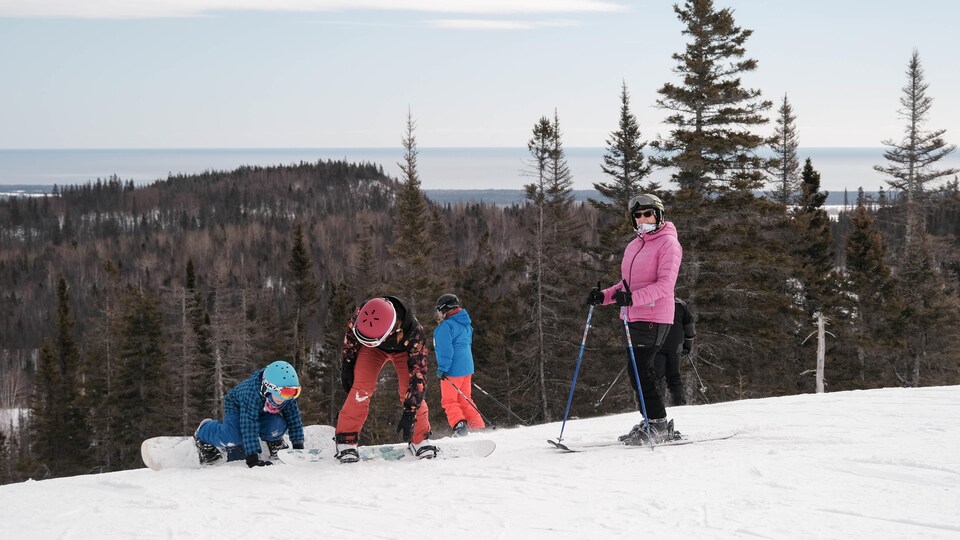 Une adulte à skis et deux jeunes en planche à neige en haut d'une pente.