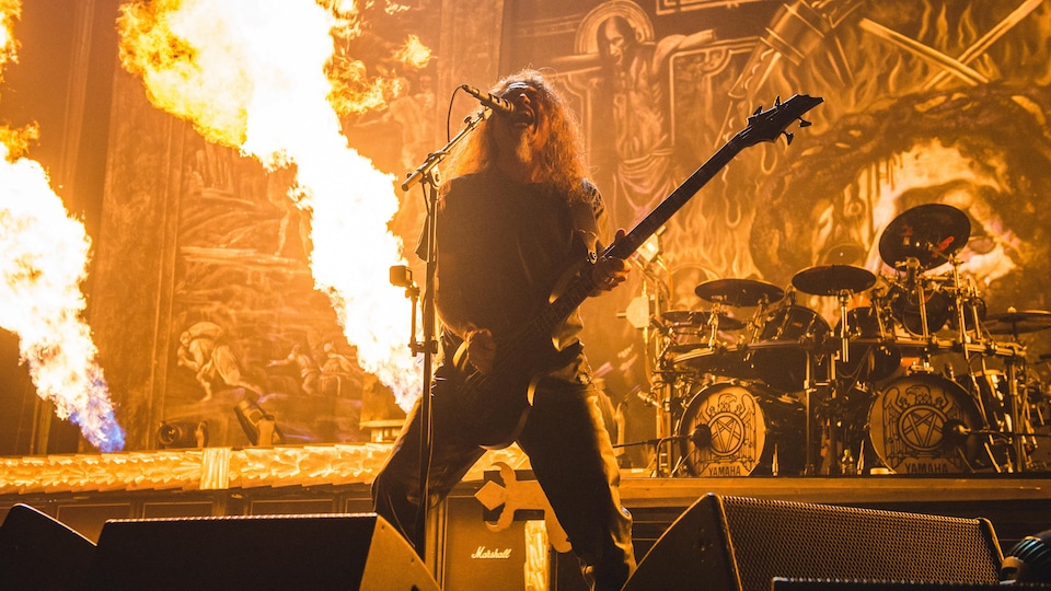 Tom Araya joue de la basse et crie dans un micro sur une scène en flammes.