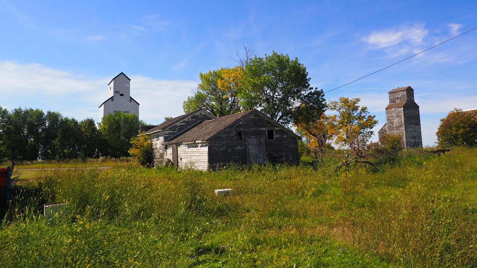 Une maison de bois abandonnée derrière laquelle on peut voir deux anciens élévateurs à grain.