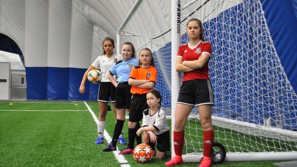 Cinq joueuses de soccer du club Greater Sudbury Soccer Club prennent la pose devant un filet de but de soccer.