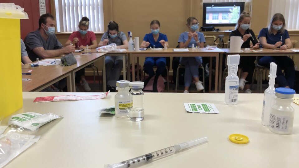 Une seringue et une fiole sur une table devant des étudiants assis à une autre table.