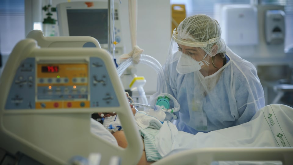 Une infirmière portant de l'équipement de protection se penche au-dessus du lit d'un patient.