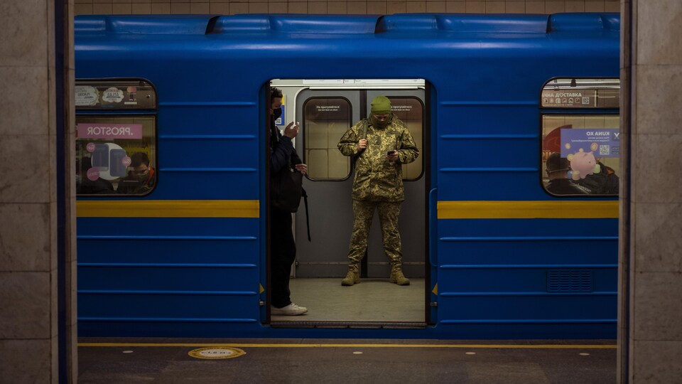 Un homme en treillis militaire consulte son téléphone. On le voit par une porte de métro ouverte sur un quai. 