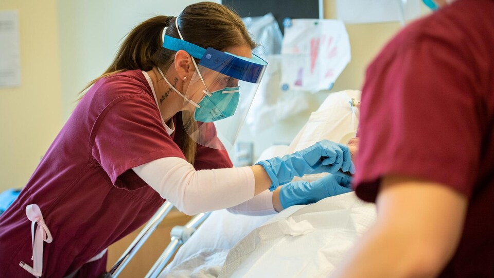 La capitaine en uniforme bourgogne porte le masque, une visière et des gants alors qu'elle apporte des soins à un patient que l'on ne peut identifier.