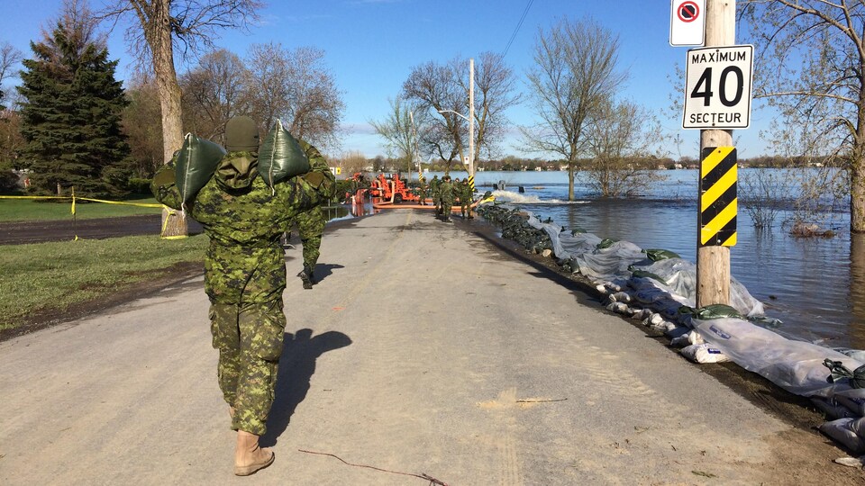 Un soldat des Forces armées canadiennes apporte des sacs de sable devant servir à renforcer une digue dans le secteur du boulevard Lalande et de la rue Rose, dans Pierrefonds-Roxboro, pendant que d'autres soldats travaillent au loin.