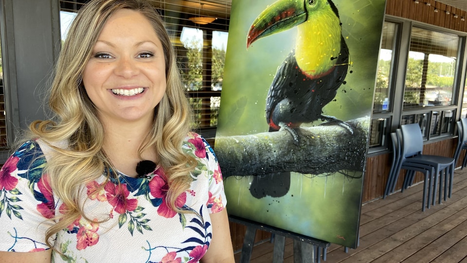 Une femme sourit devant une toile d'un toucan.