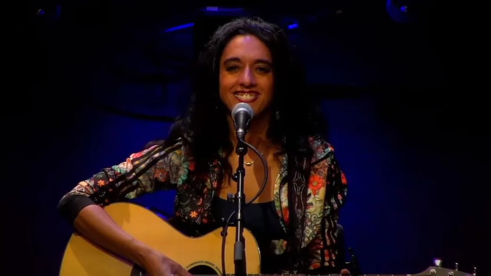 Une femme assise avec une guitare dans les bras, devant un micro.