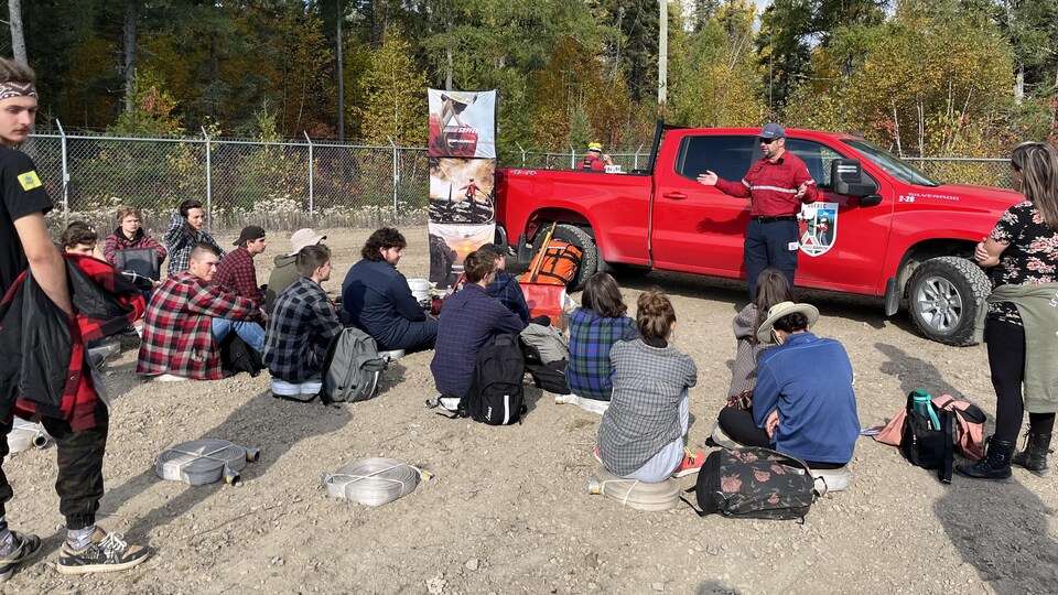 Des jeunes sont assis devant un homme qui parle devant une camionnette rouge.