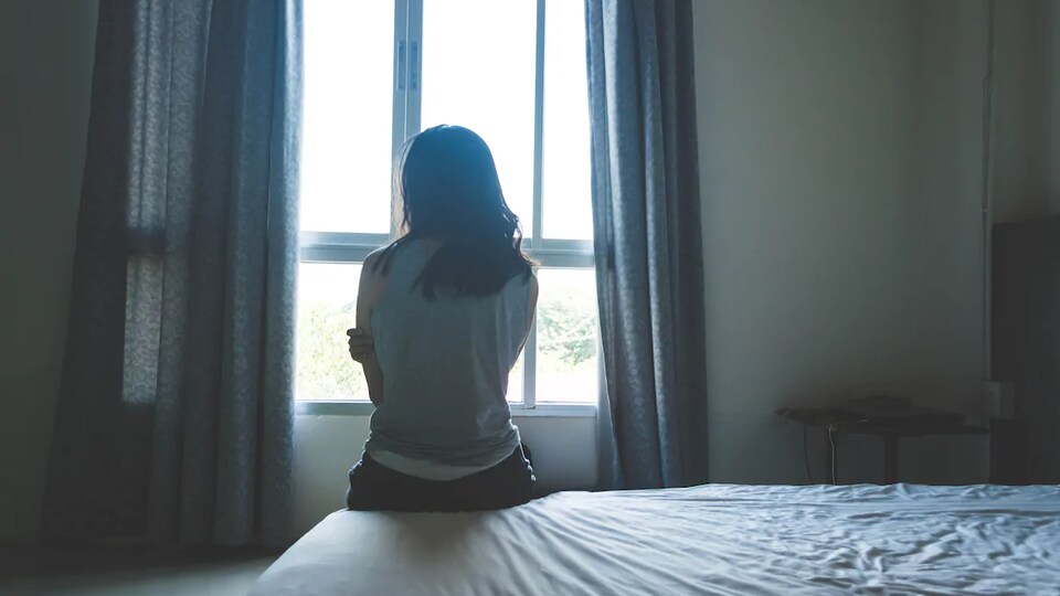 Une femme est assise sur un lit et regarde la fenêtre.