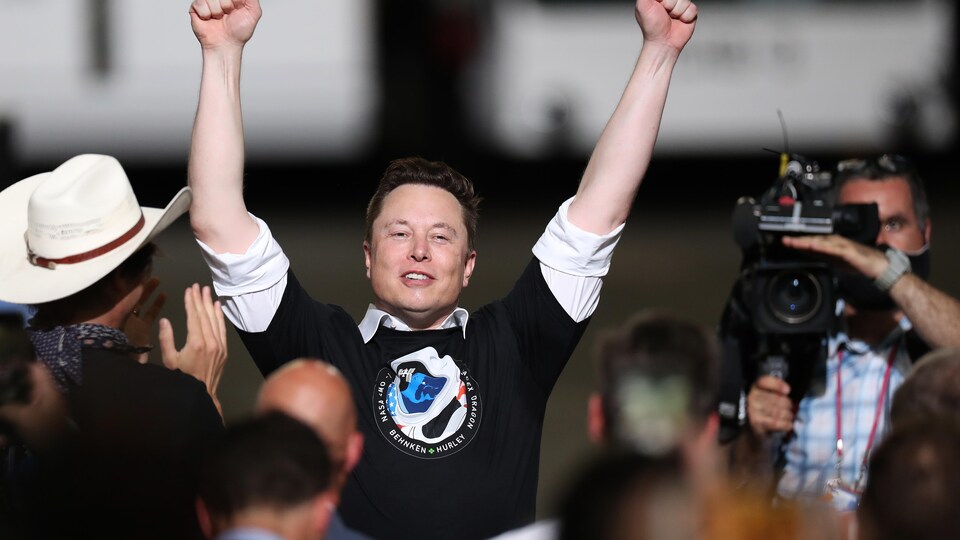 L'homme lève les bras dans les airs en signe de victoire, entouré de personnes qui applaudissent et d'un caméraman qui filme le public.