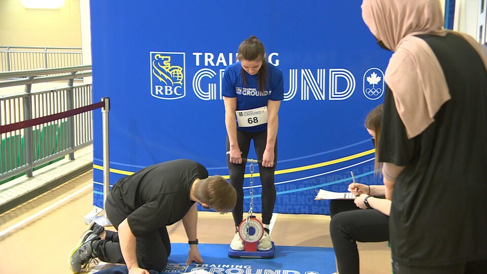 Un évaluateur à genoux mesure les données inscrites aux pieds d'une jeune athlète. 