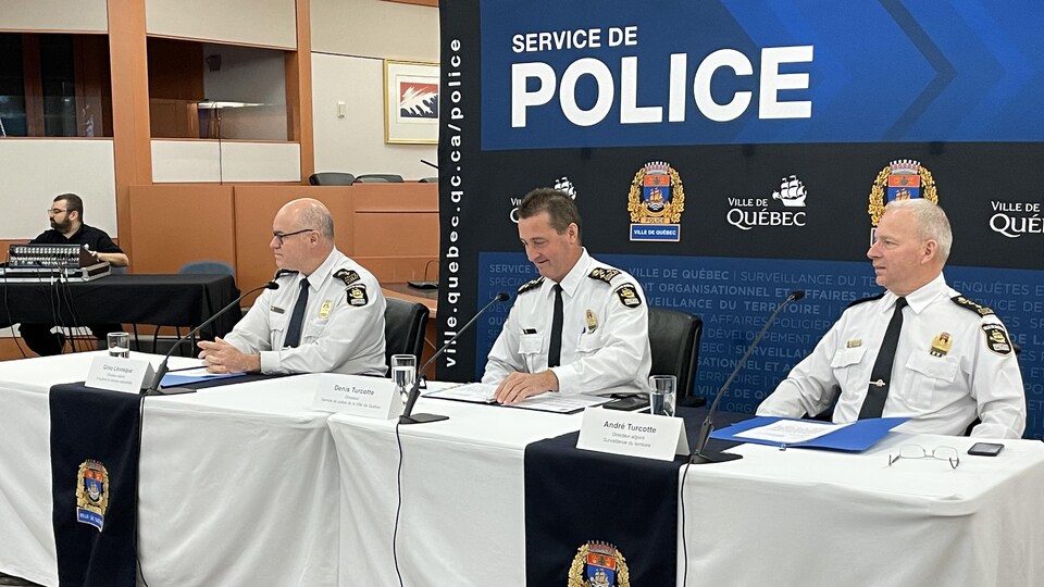 Trois hommes en uniforme du Service de police de la Ville de Québec sont assis derrière une table de conférence de presse.
