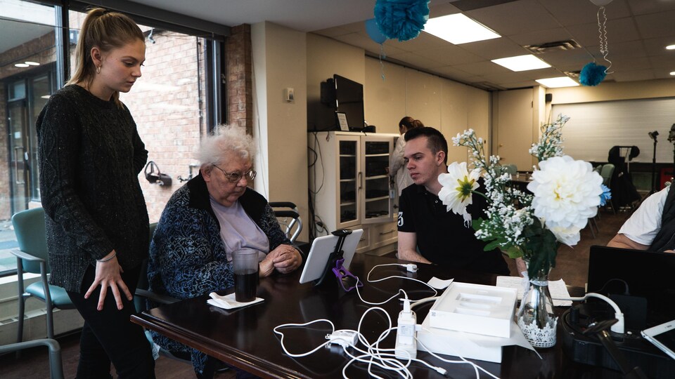 Une jeune femme et un jeune homme donne des conseils à une dame âgée assise devant une tablette. 
