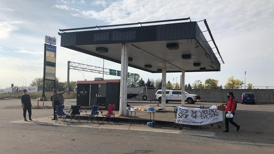 Une station-service désaffectée, occupée par quelques personnes, avec des tables, des chaises et un panneau qui dit, en anglais : « Arrêtons la violence et les expulsions ».