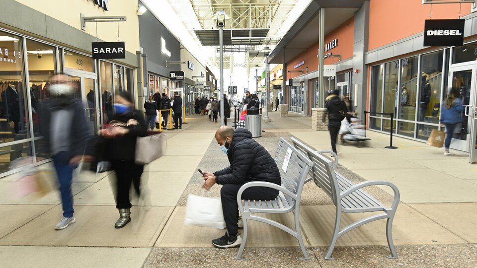 Des consommateurs déambulent dans l'allée d'un centre commercial et un homme regarde son téléphone intelligent, assis sur un banc.