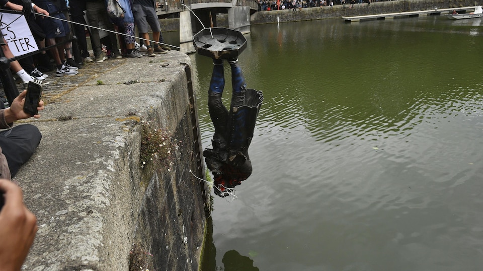 Une statue, tête en bas, en retenue par des cordes par des manifestants. Ils s'apprêtent à la jeter dans une rivière.