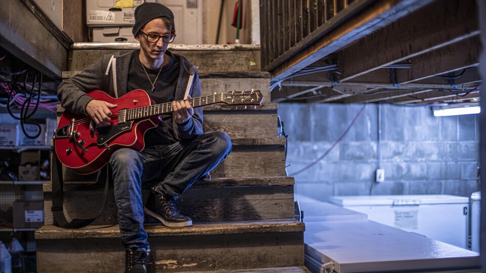 Assis dans les marches d'un escalier de bois, un homme gratte les cordes de sa guitare rouge.