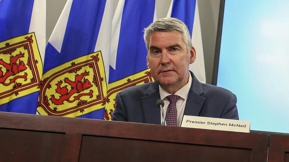 Le premier ministre assis à une table, avec son nom écrit devant lui, devant des drapeaux de la Nouvelle-Écosse.
