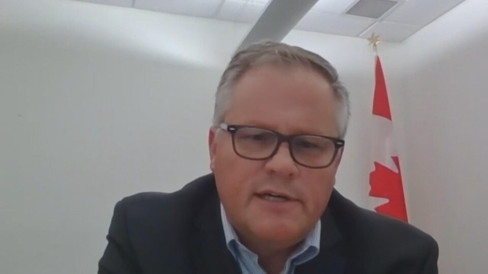 Un homme avec des lunettes et un costume, un drapeau canadien derrière lui.