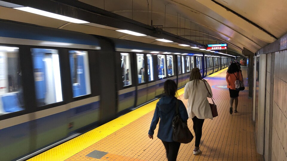 Des gens marchent sur le quai du métro, tandis qu'un train passe.
 
