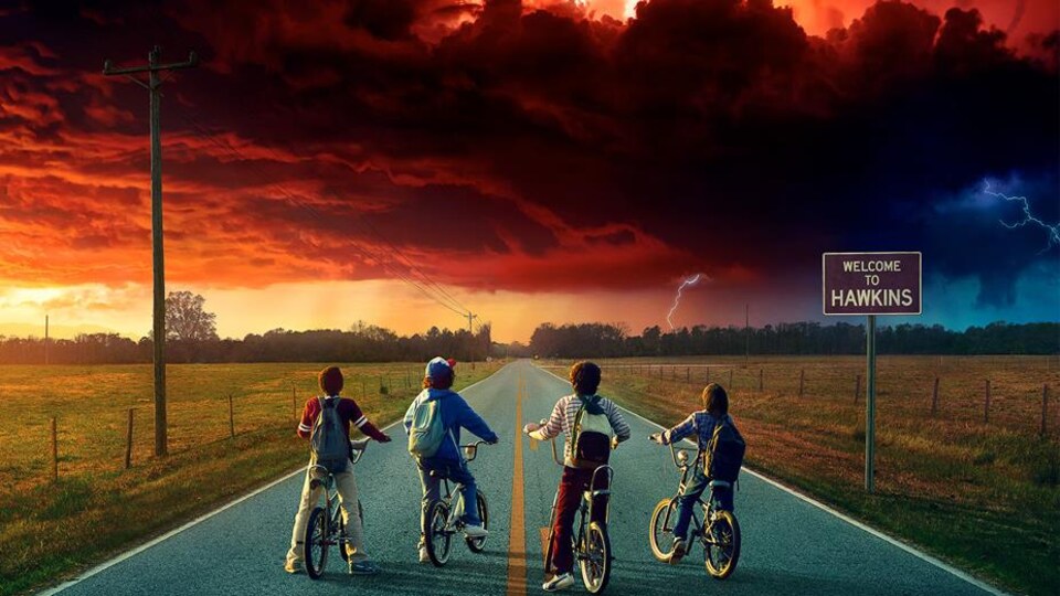 Quatre adolescents immobilisés sur leurs bicyclettes se trouvent sur une longue route et regardent un ciel 
 bondé de nuages rouges. 