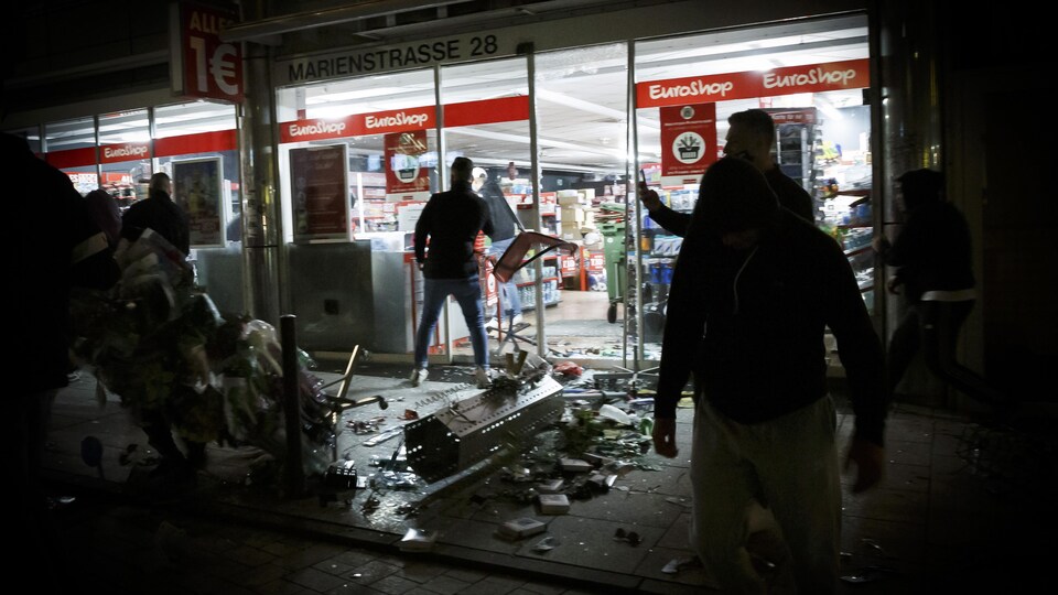 Des produits d'un magasin vandalisé jonchent le sol, dans la nuit, tandis que des gens observent la scène dans la pénombre.