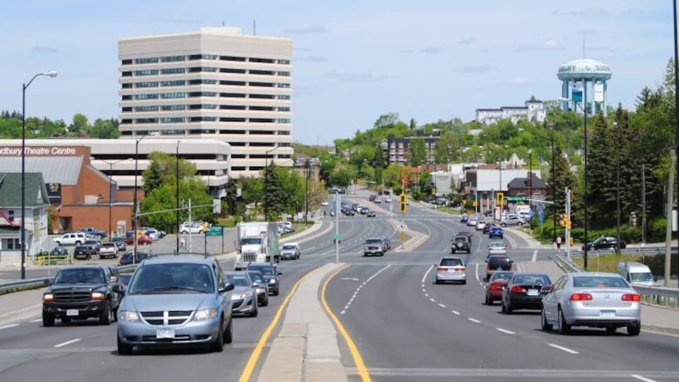 Une rue du centre-ville de Sudbury. Les voitures sont en mouvement. Au loin, le château d'eau de Sudbury.