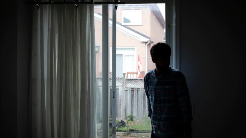 Un homme non identifiable se tient dans l'ombre, près d'une fenêtre.
