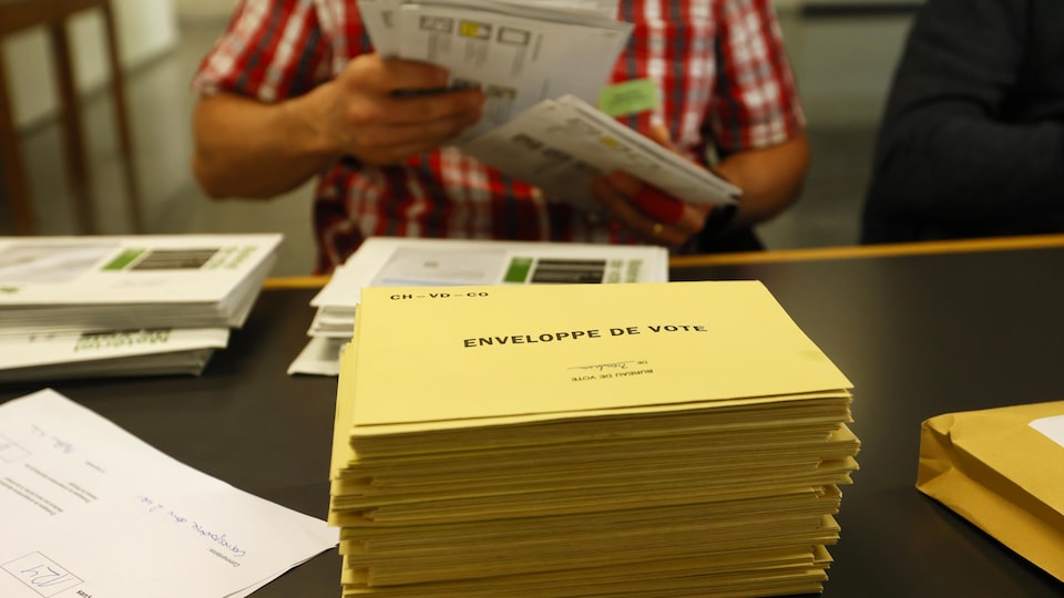 Une pile d'enveloppes de vote sur une table. Un homme en tient quelques-unes dans ses mains. 