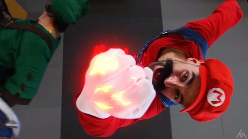 Une capture d'écran de la vidéo montrant un homme déguisé en Super Mario, avec sa salopette bleue, son chandail et sa casquette rouge et sa moustache, en train de brandir son poing en feu devant la caméra.