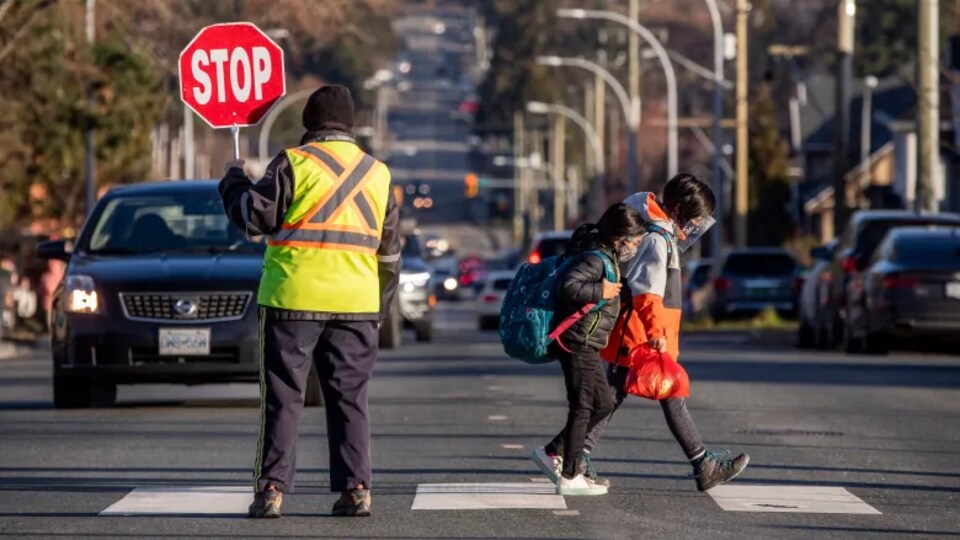 Deux enfants traversent un passage pour piétons pendant qu'un agent tenant un panneau d'arrêt stoppe la circulation routière.