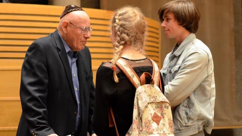 Le survivant de l'Holocauste Janos Benisz est debout derrière les chaises d'un auditorium et parle à deux jeunes étudiants qui l'écoutent avec attention.