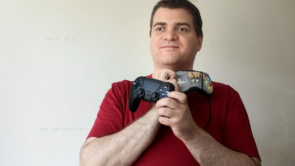 Portrait de l'homme qui tient sa manette d'une main et son masque de l'autre devant un mur blanc.