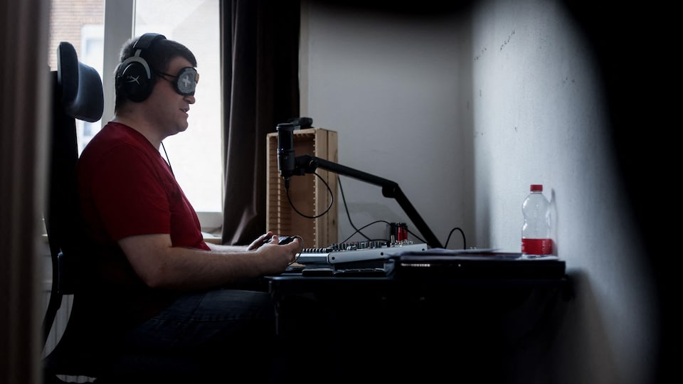 L'homme avec un bandeau sur les yeux et des écouteurs, tient une console et est assis à son bureau devant un mur blanc sans écran.