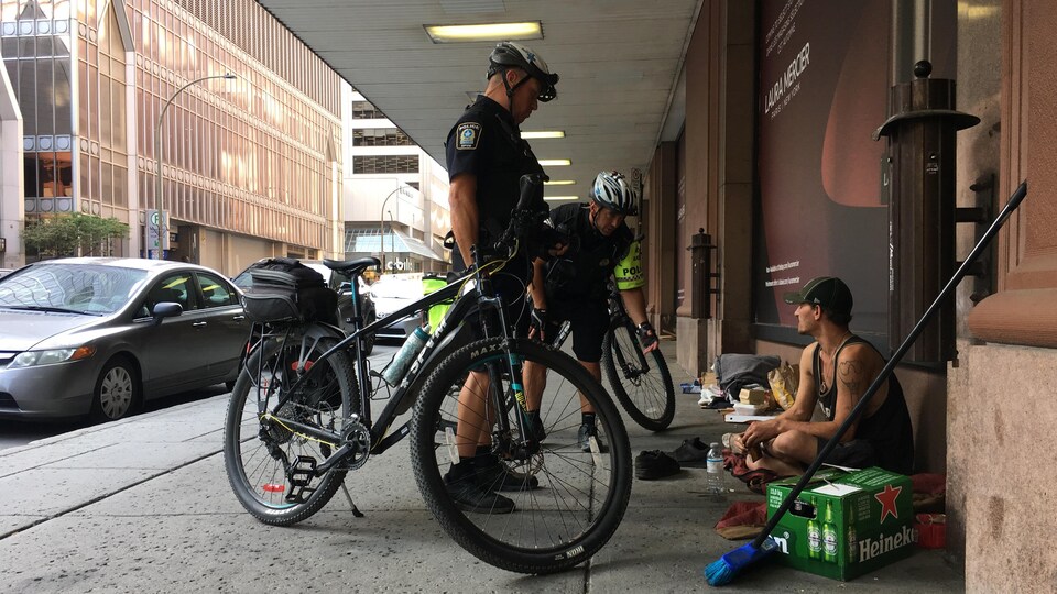 Deux policiers près de leur vélo discutent avec un itinérant assis sur le trottoir.