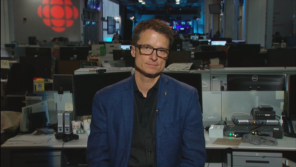 Un homme est assis dans une salle de presse. À sa droite se trouve le logo de Radio-Canada.