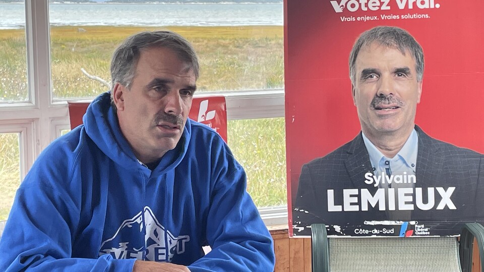 Un homme est assis devant une fenêtre; une pancarte électorale avec sa photo est à côté de lui.