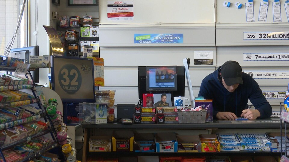 Un homme est accoudé au comptoir de son dépanneur. Il note quelque chose sur un calepin.