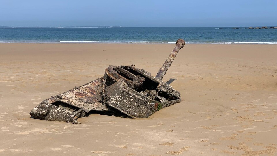 Un char d'assaut rouillé partiellement enfoui dans le sable sur une plage.