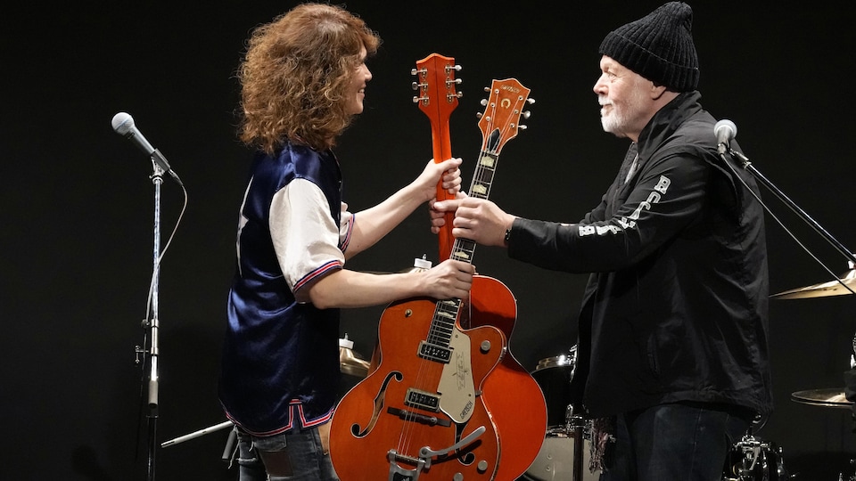 Face à face sur une scène, deux personnes s'échangent deux guitares.