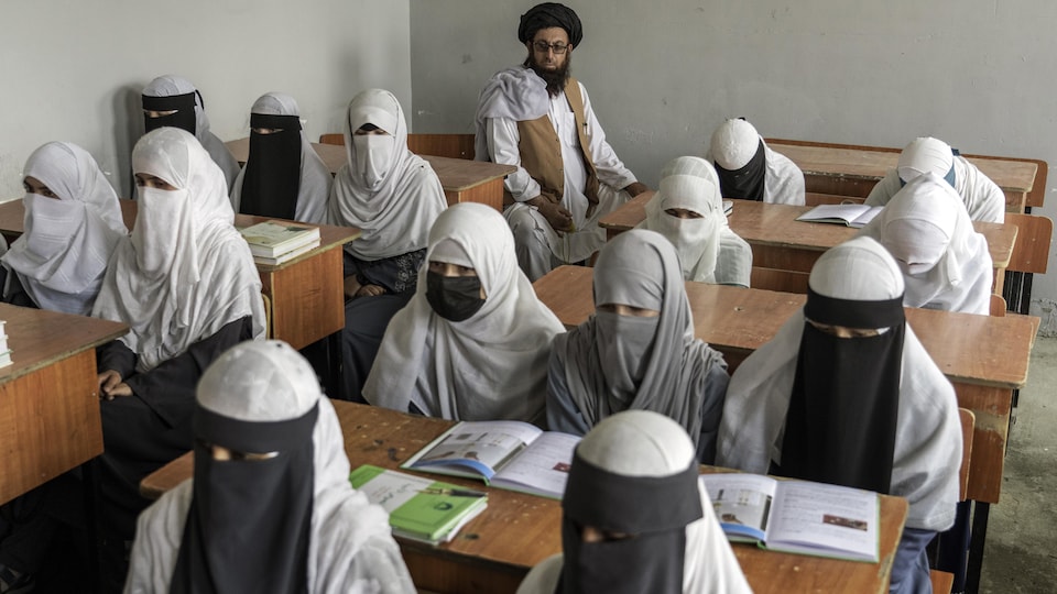 Des femmes portant la burqa, assises derrière des pupitres de bois, surveillées par un homme au fond de la classe.