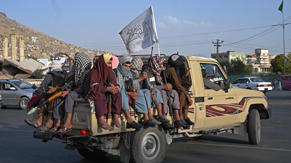 Plusieurs hommes armées sont assis dans la boîte d'une camionnette, surmontée d'un drapeau des talibans. 